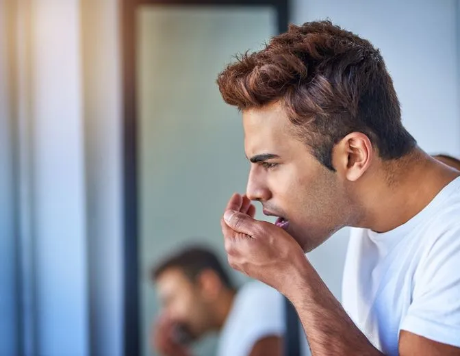 A man is covering his mouth while looking at himself in the mirror.